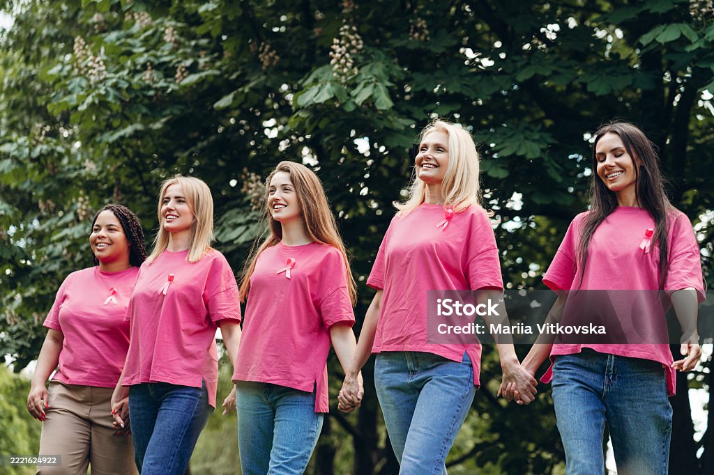 Women of mixed age range are holding hands and walking together in park during breast cancer awareness walk